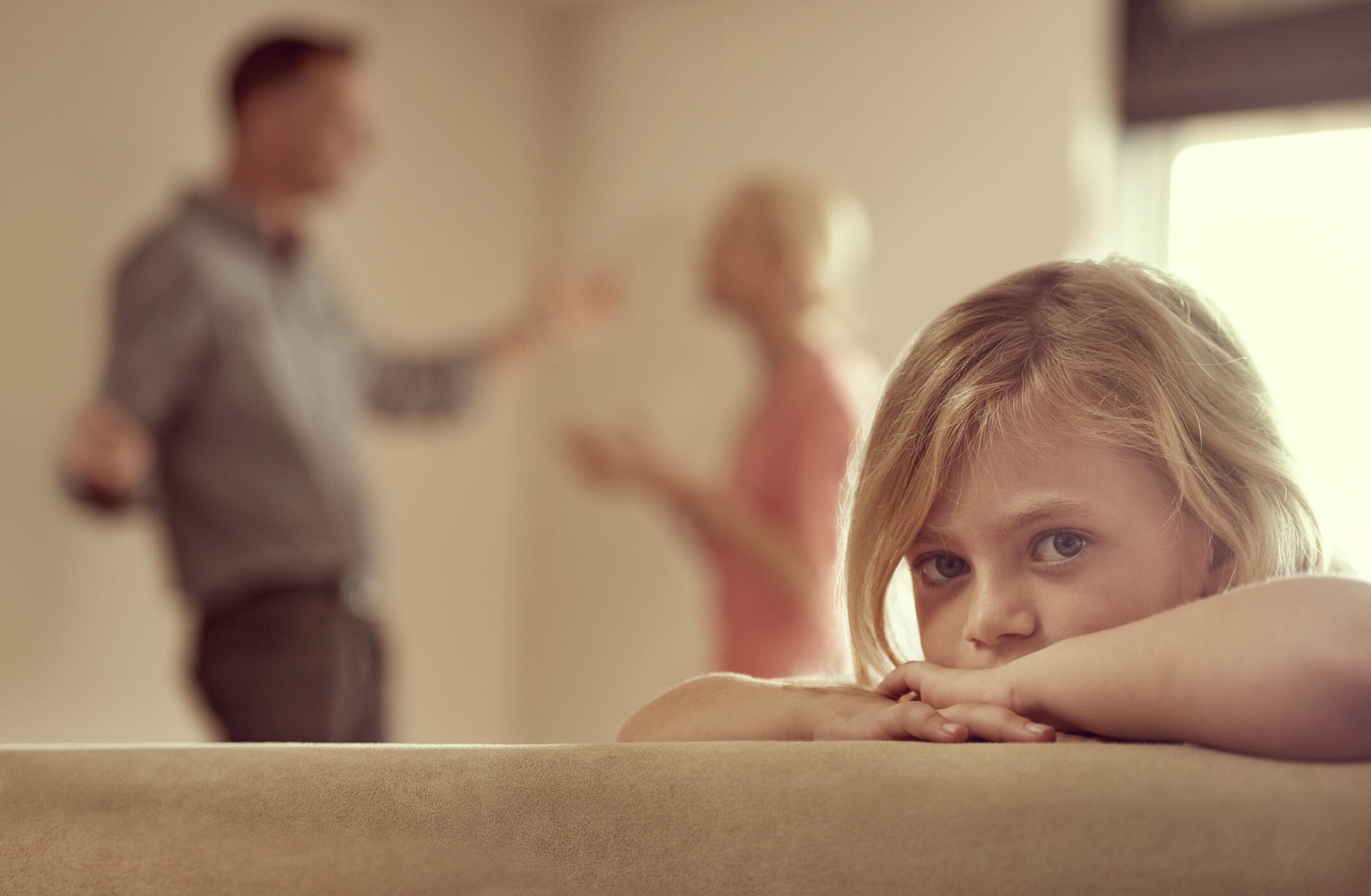 A child sadly leaning on a couch while in the background a blurry image of a woman and man look like they're fighting with each other. 