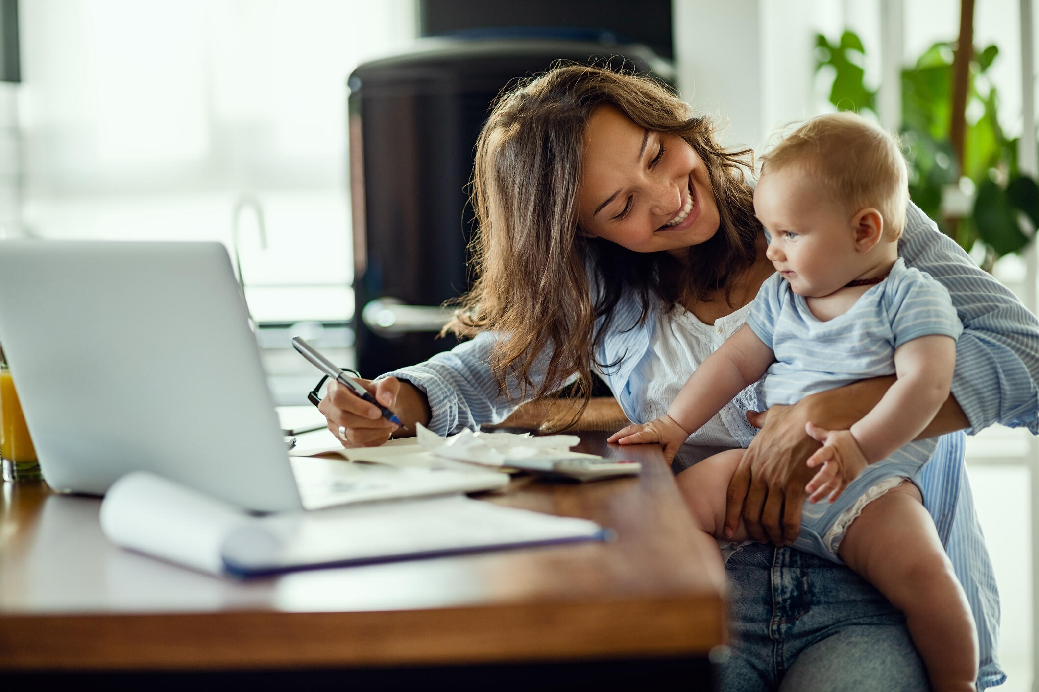 A woman holding a baby and smiling while sitting in front of a laptop. 