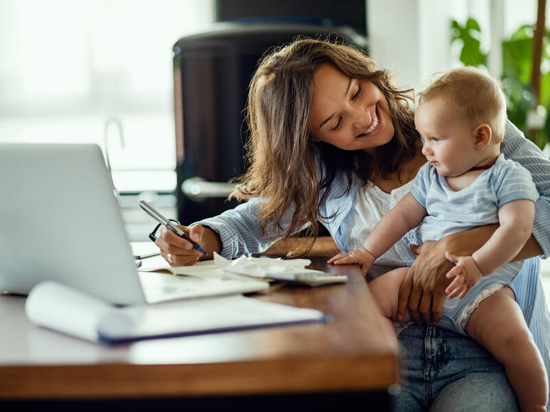 A woman holding a baby and smiling while sitting in front of a laptop. 
