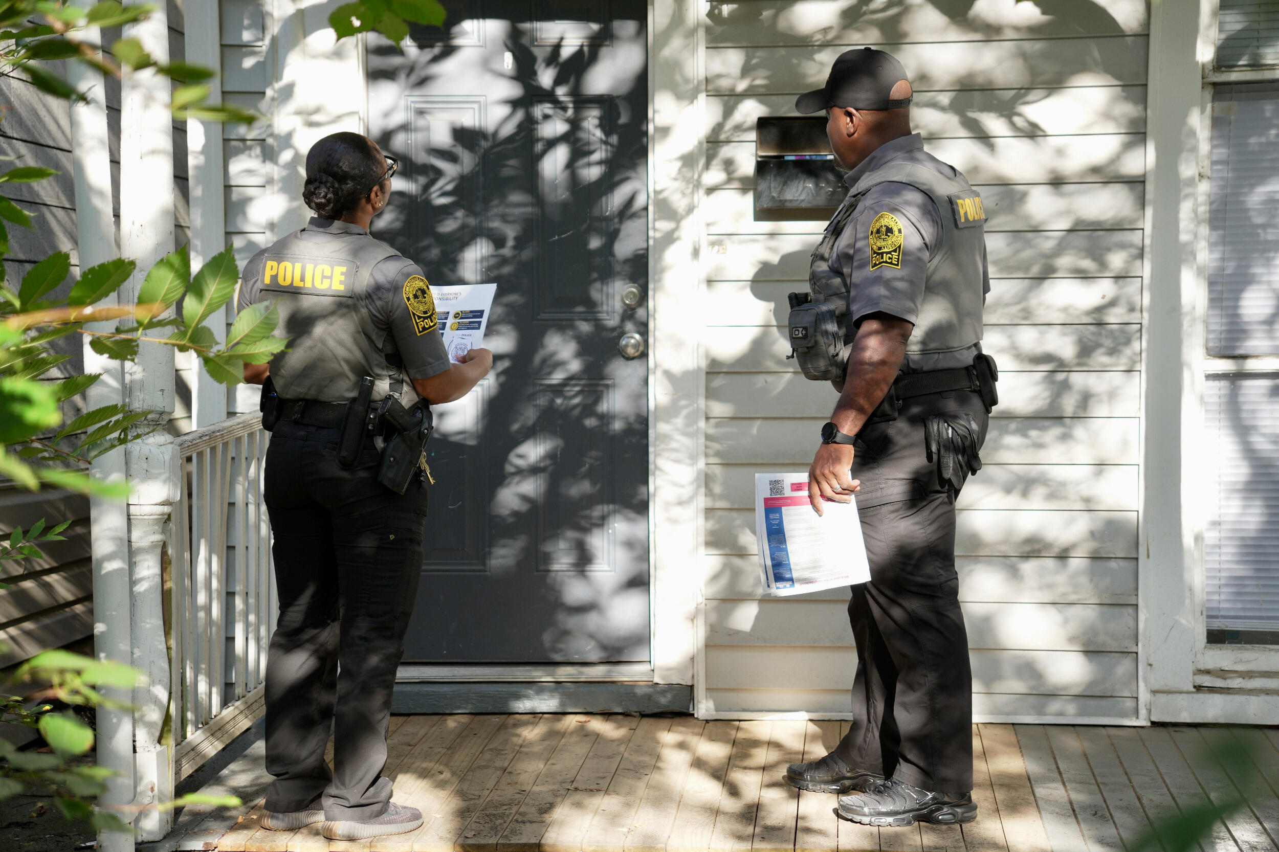 Two police officers holding sheets of paper wait at the door of a residence.
