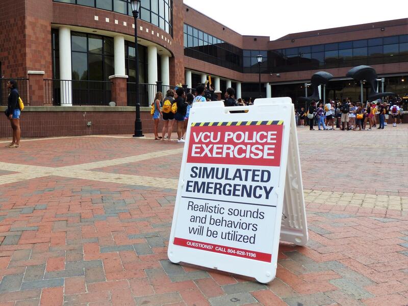 A photo of a sign on the ground in the commons plaza that says \"VCU POLICE EXERCISE SIMULATED EMERGENCY\" \"Realistic sounds and behaviors will be utalized.\" Behind the sign, a few groups of people in civilian clother are standing and talking to each other. 