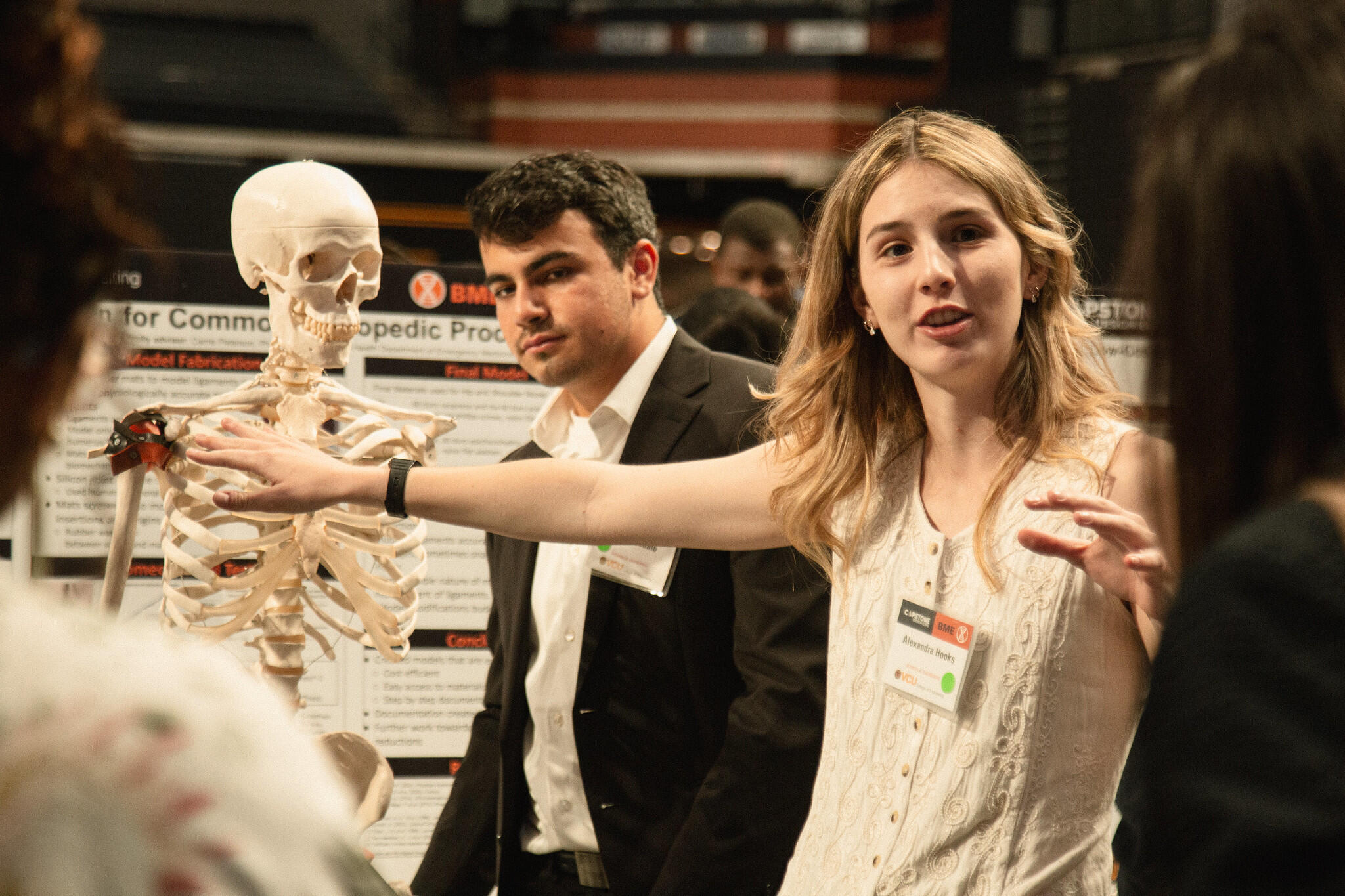 A photo of a woman speaking and pointing at a model skeleton. There are two people in front of the woman who are listening to her speak. There is a man behind the woman who is looking at the other two people. 