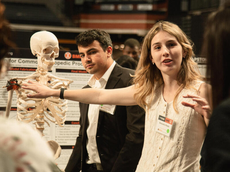 A photo of a woman speaking and pointing at a model skeleton. There are two people in front of the woman who are listening to her speak. There is a man behind the woman who is looking at the other two people. 