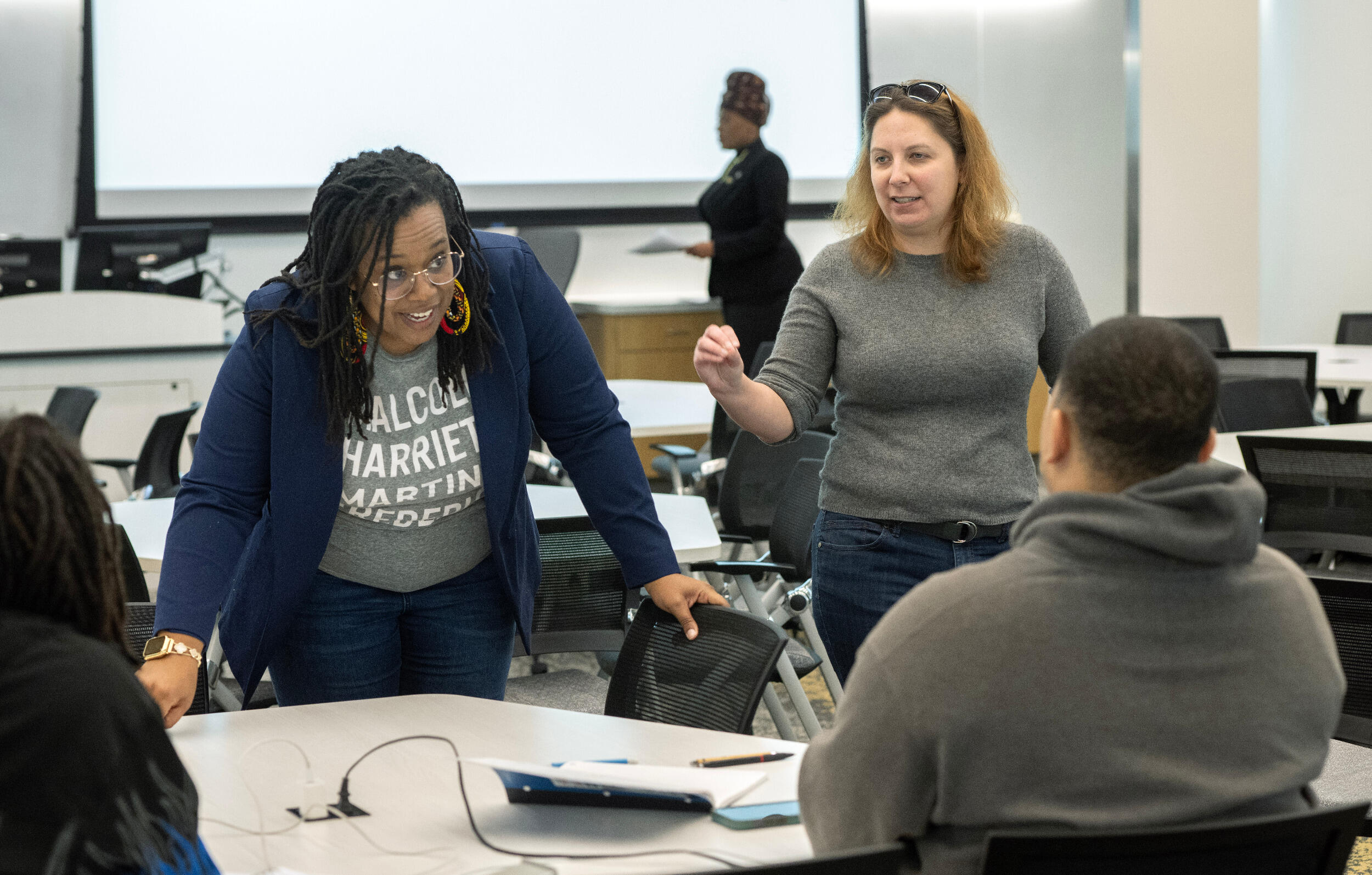 Two teachers stand while speaking with seated students.