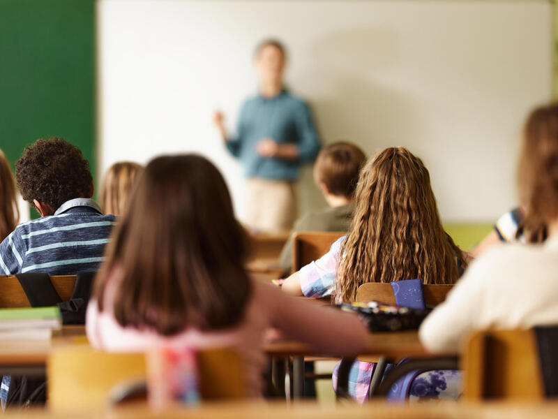 Children at desks in a classroom watch a teacher at the front of room talking.