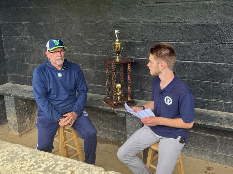 A photo of two men sitting on benches. The man on the right has a stack of paper in his hands. Behind the two men is a large trophy. 