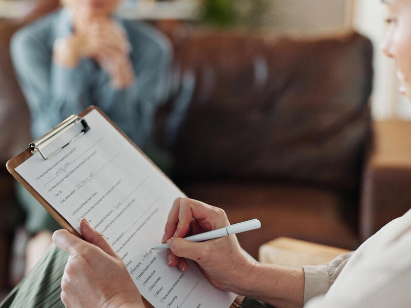 A photo of two people sitting in a room from across from eachother. One person is sittin on a couch, and the other is sitting in a chair and holding a pen and clip board. 