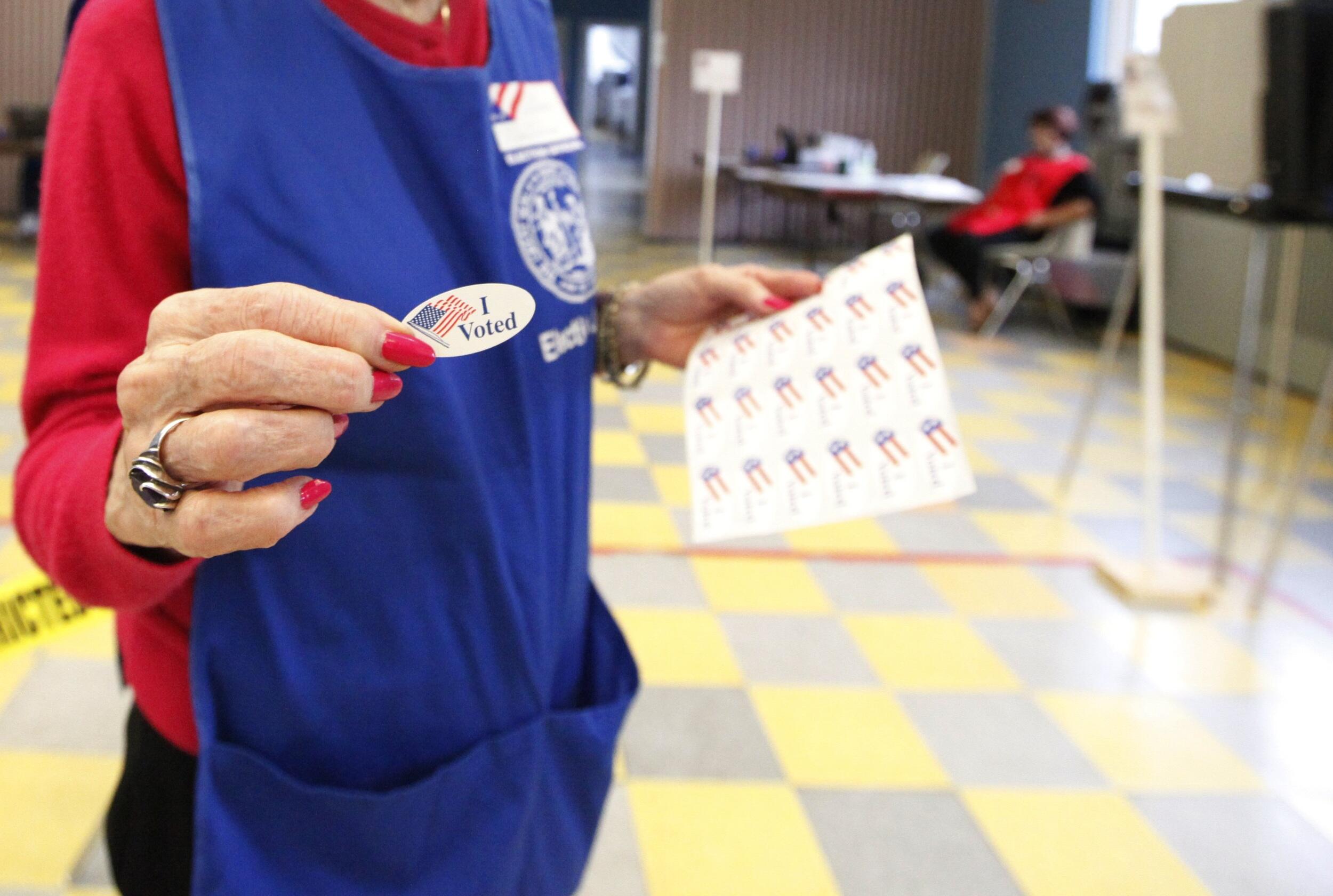 A person stands holding a sheet of stickers in one hand and an \"I voted\" sticker in the other.