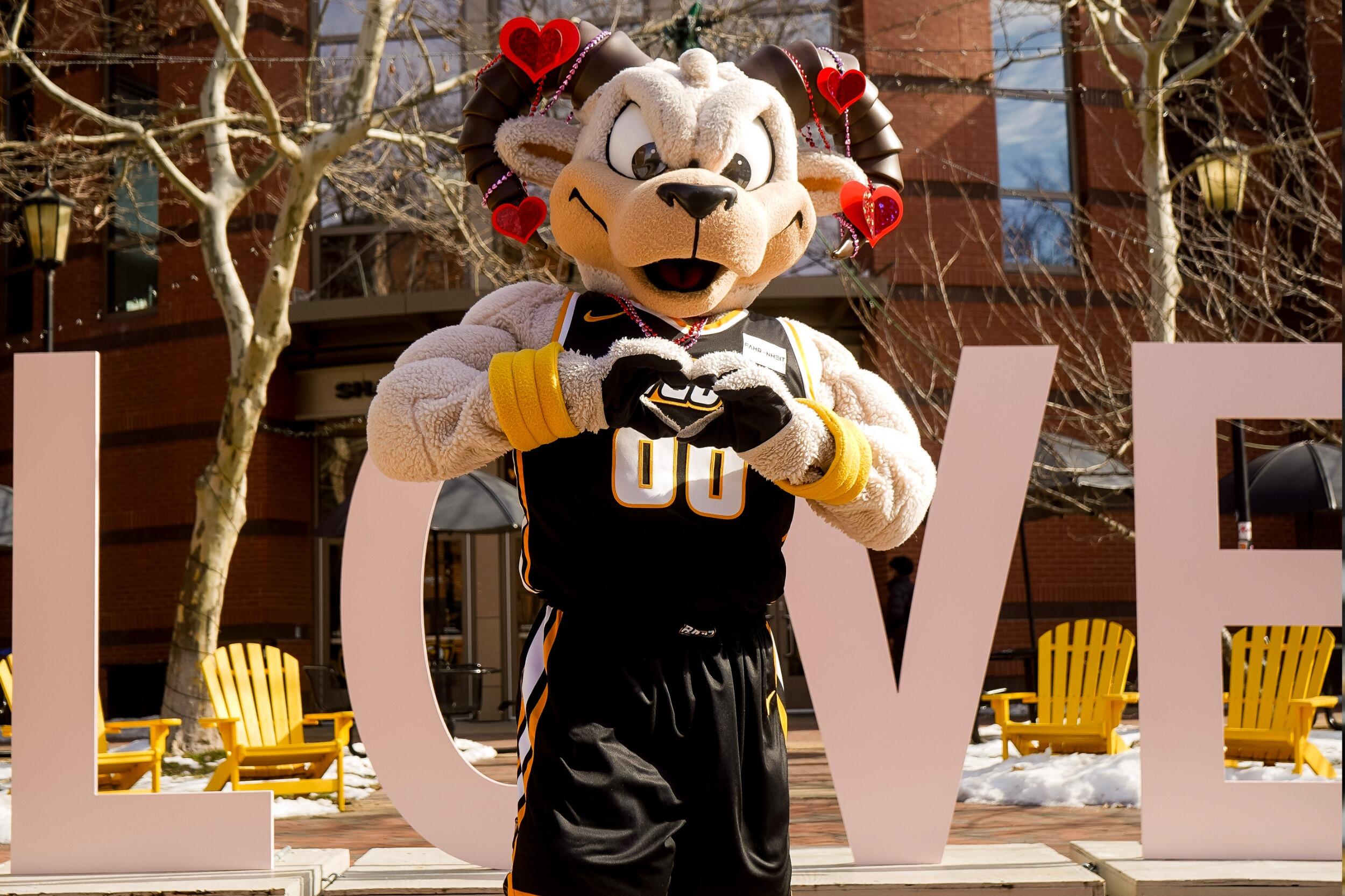 Mascot Rodney the Ram makes a heart shape with his hands, and stands in front of giant letters that spell love. His horns are decorated with hearts.
