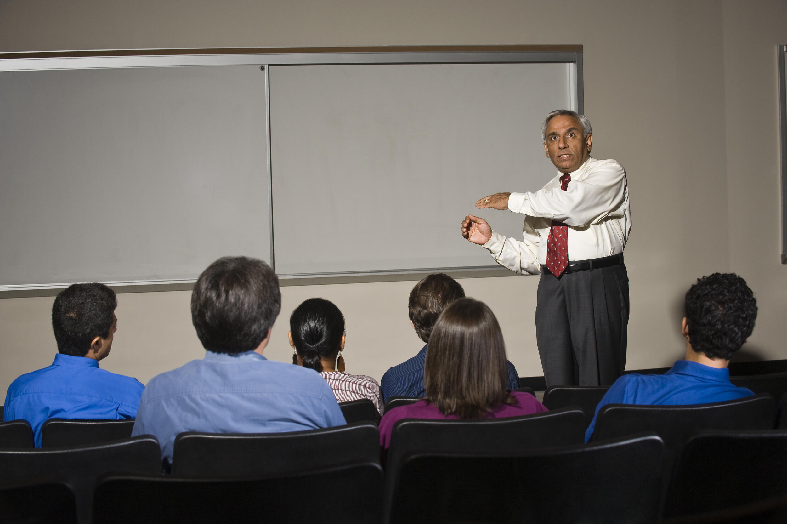 A photo of a man standing at the front of a classroom and motioning towards a white board while talking to students. 