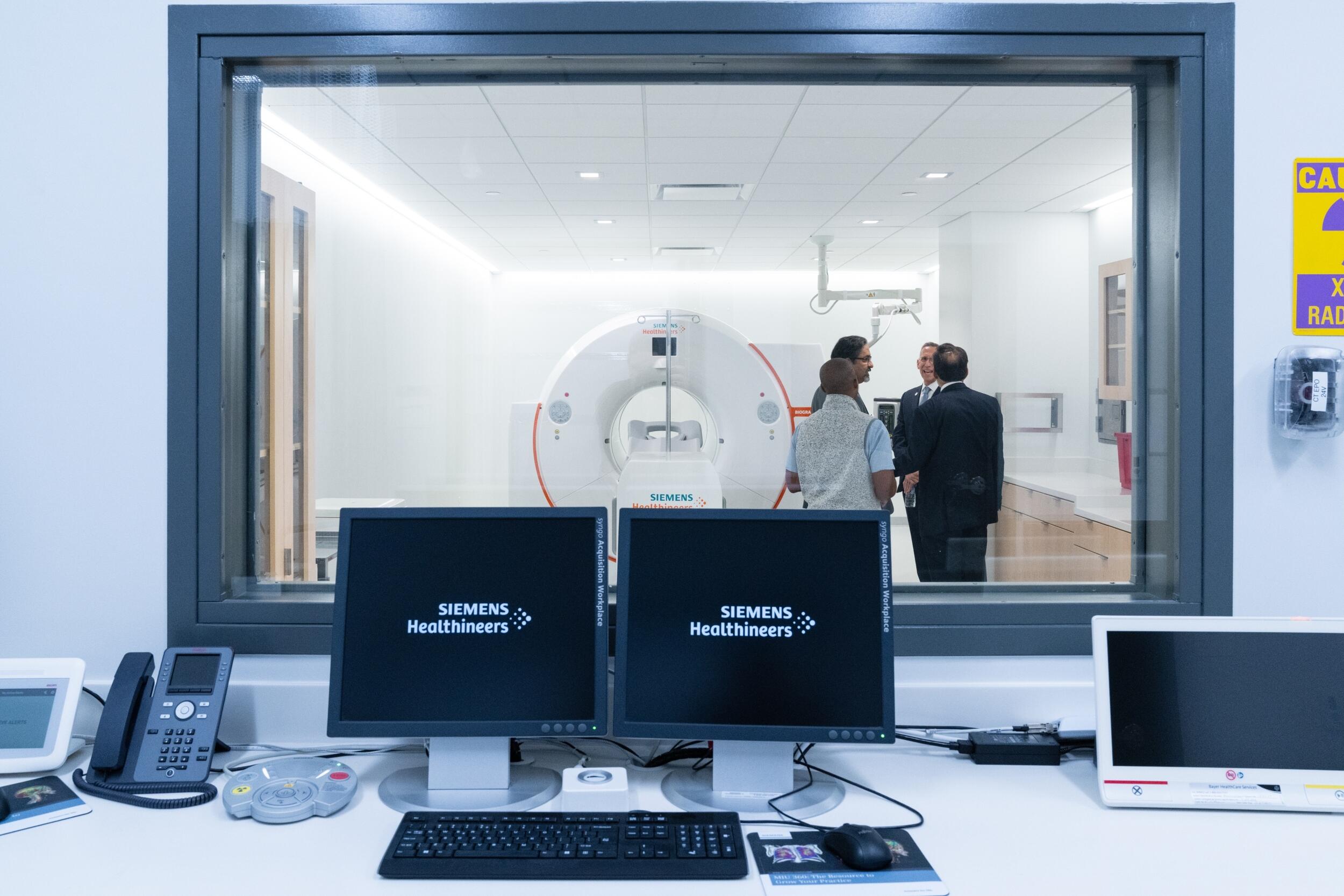 A photo of a room with a window that shows a PET/CT scanner. 