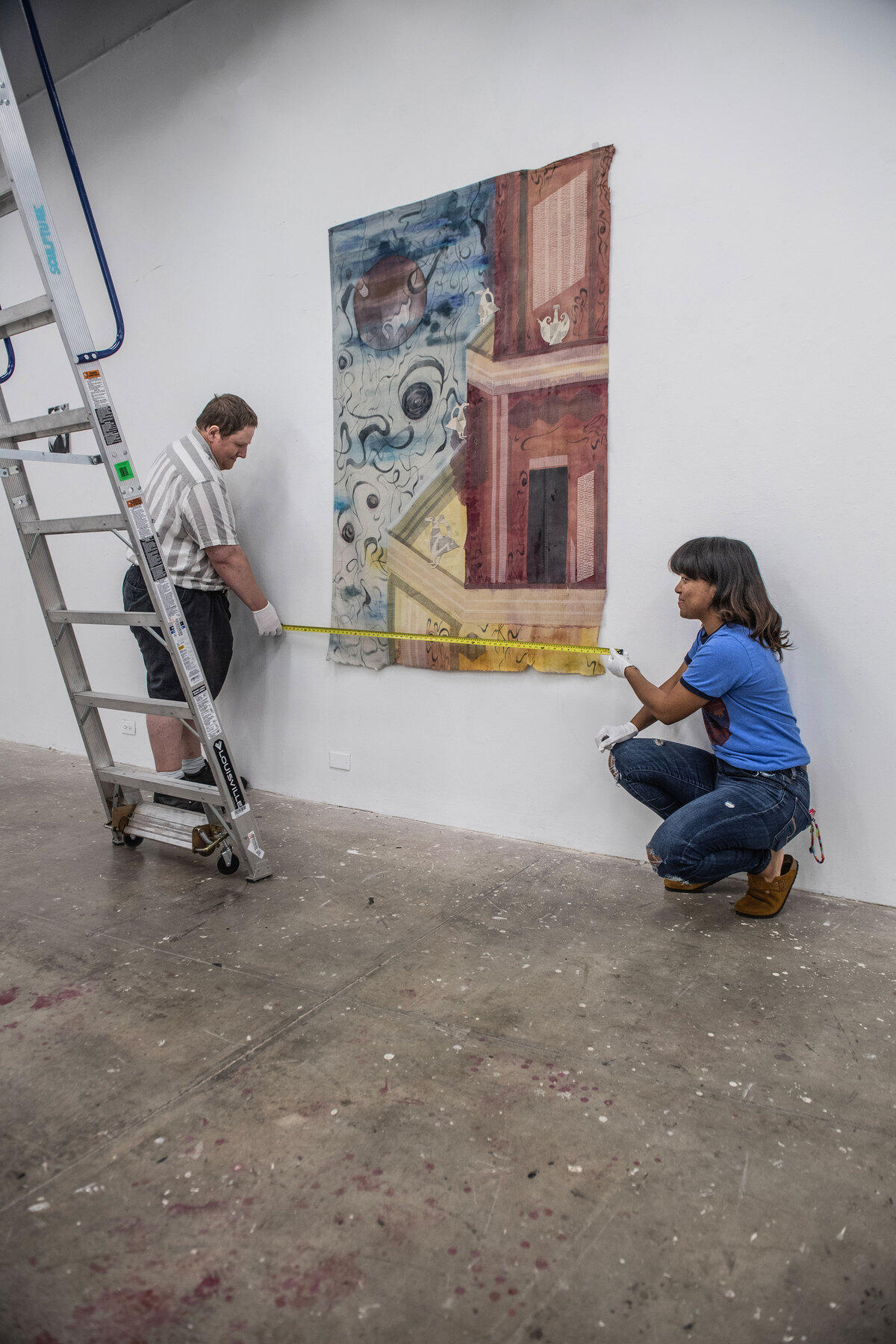 A man is standing to the left of a painting and a woman is kneeling to the right of it. They are both holding measuring tape going across the painting. 