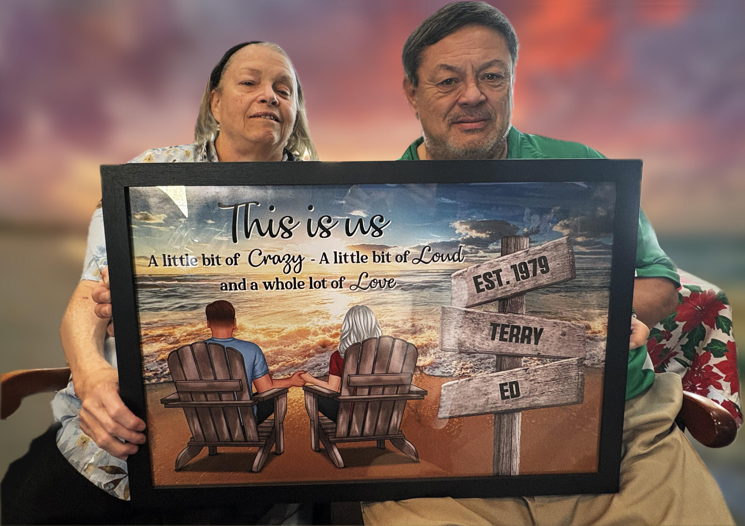 A woman and a man hold a painting in front of them of a man and woman sitting in chairs on the beach.