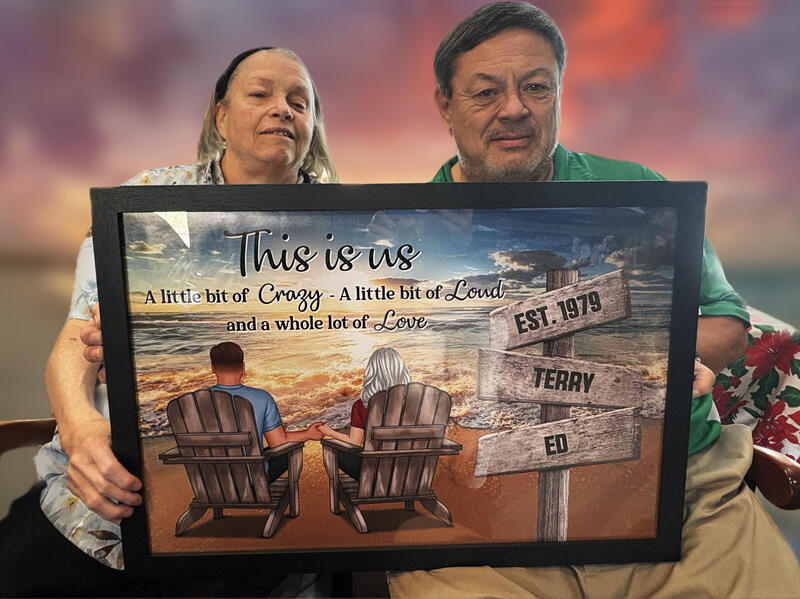 A woman and a man hold a painting in front of them of a man and woman sitting in chairs on the beach.