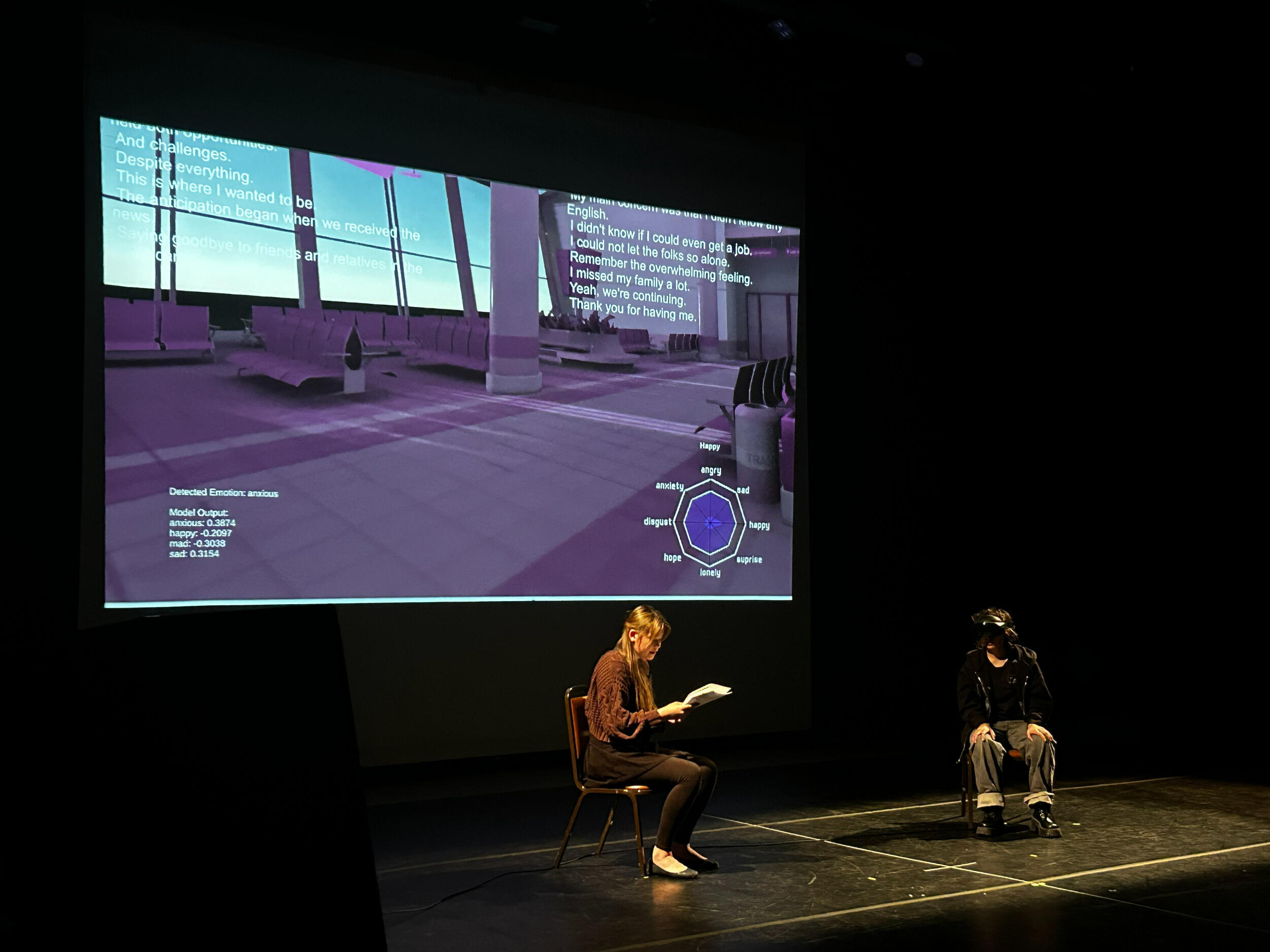 A photo of two people sitting in chairs on a stage. The person on the left is holding a stack of papers they are reading from and the person on the left is wearing a VR headset. Behind them is a large screen that shows a 3D image of what looks like an airport waiting area.