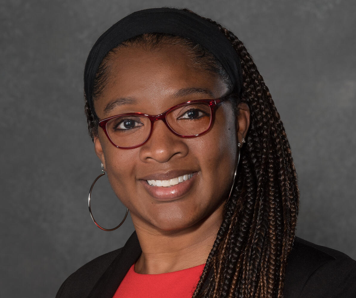 A portrait of a woman with long braids wearing a black cardigan, glasses, and hoop earrings. 