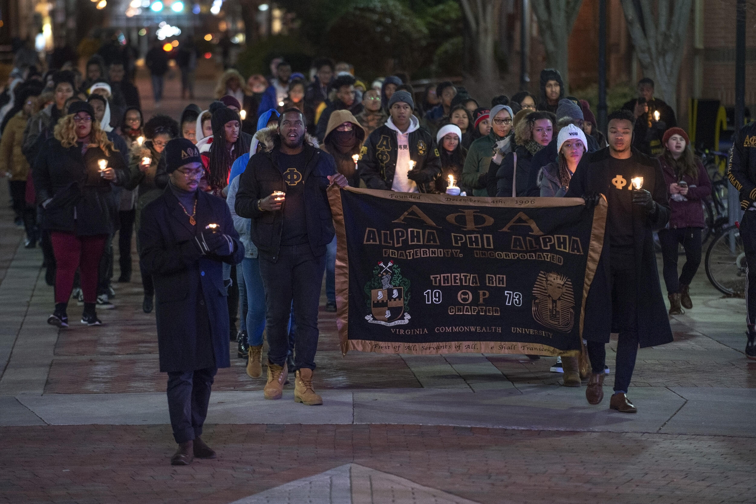 A photo of a group of people walking with candles. Two people in the front of the group hold up a banner for Alpha Phi Alpha. 