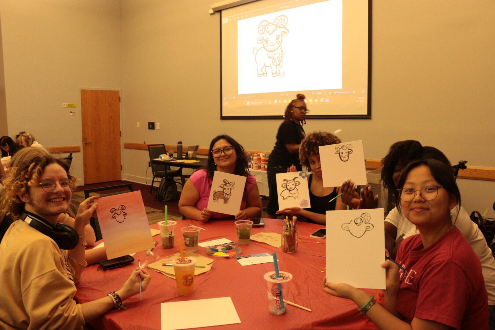 A photo of five people sitting at a table holding up illustrations of rams they drew. 