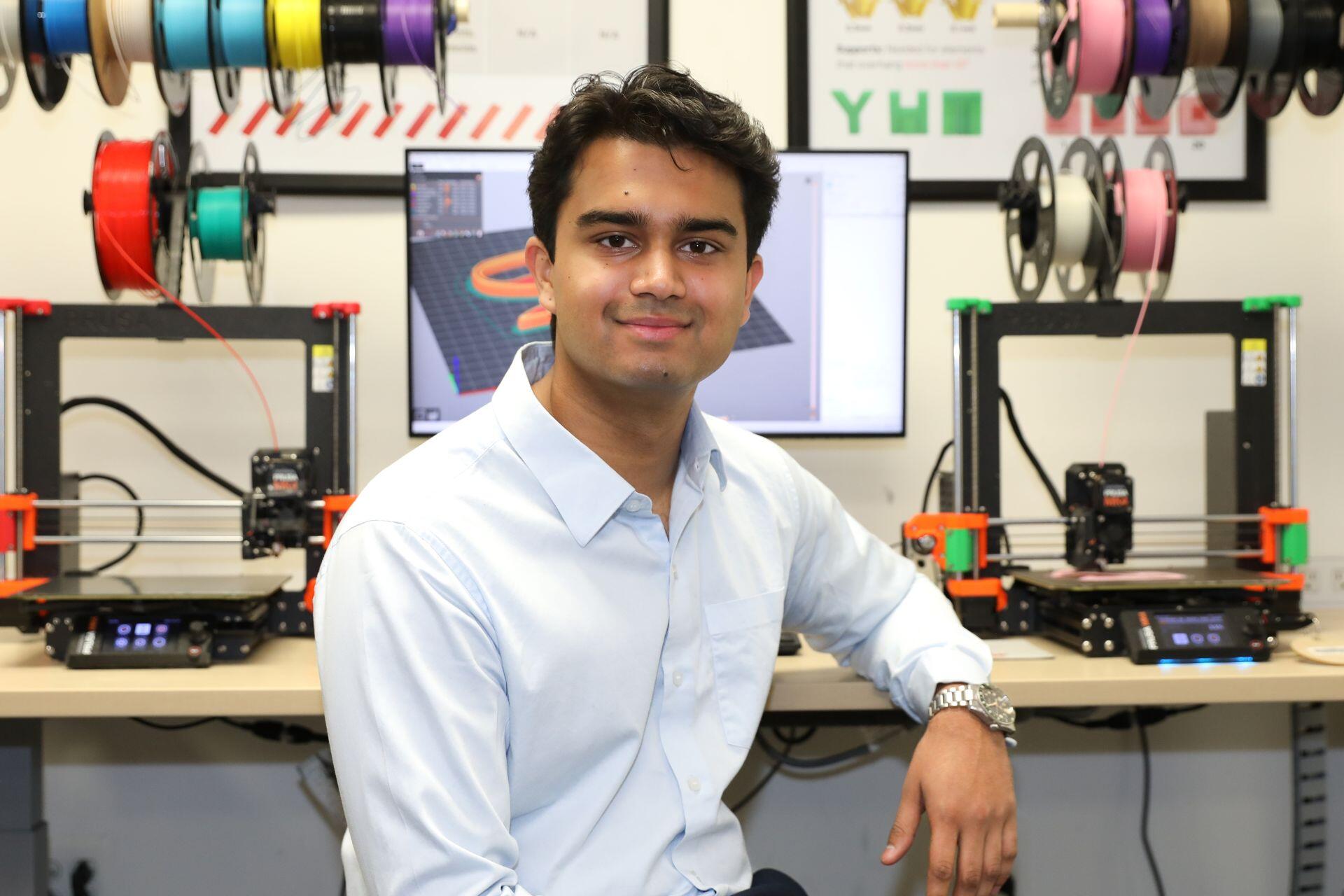 A photo of a man sitting at a desk with a computer screen and two 3D printers on it. 