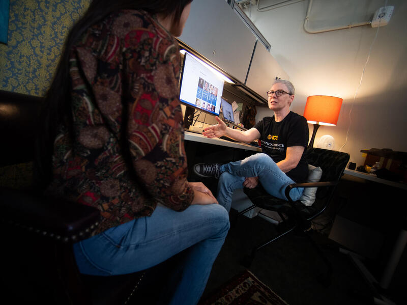 A photo of a woman seated at a desk talking to a woman who is sitting in a chair across from it. 