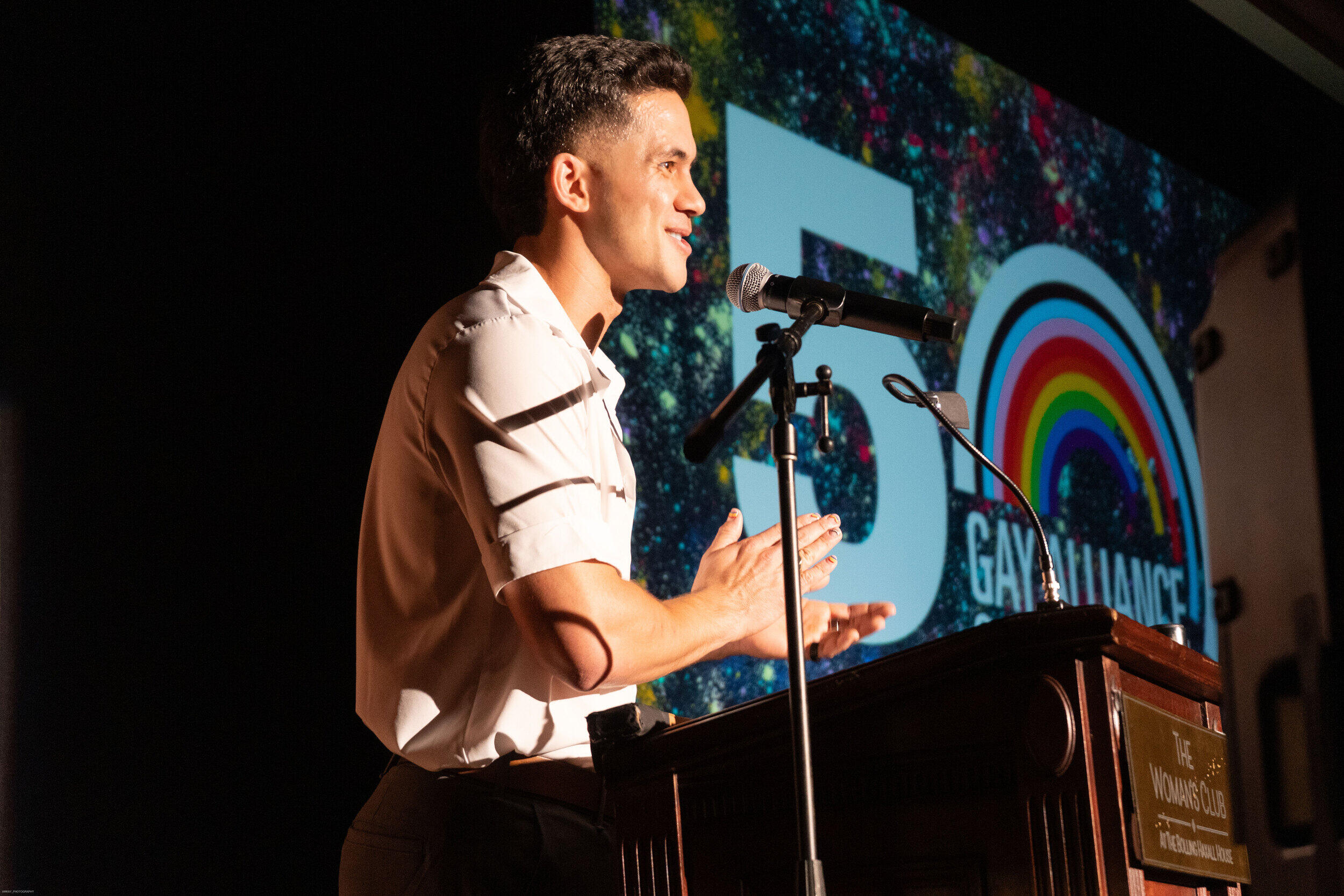 A photo of a man standing behind a podium. Next to him is a screen that says \"50\" \"Gay Alliance's\"