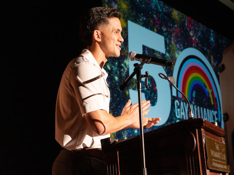 A photo of a man standing behind a podium. Next to him is a screen that says \"50\" \"Gay Alliance's\"