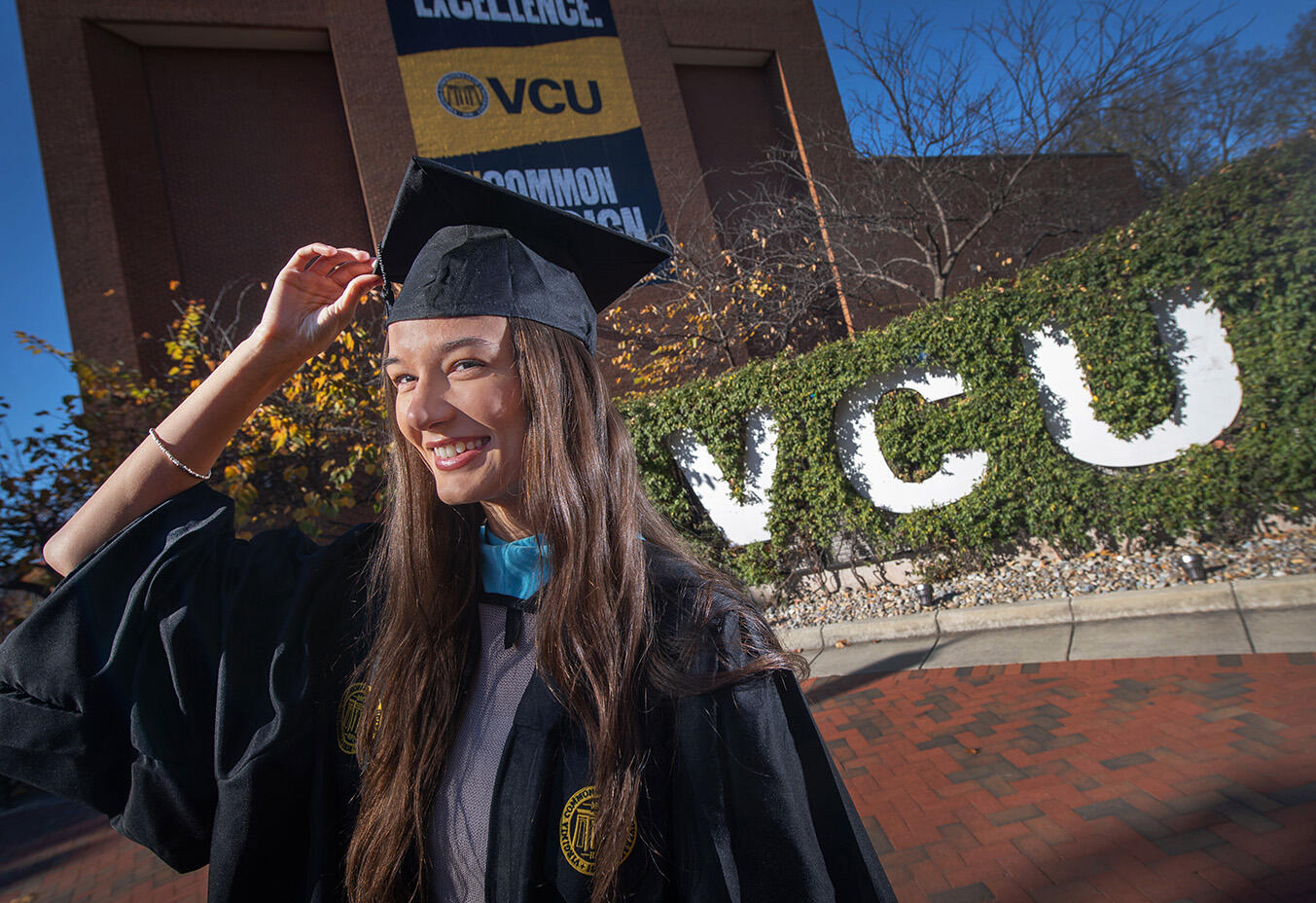 A photo of a woman wearing a graduation cap and gown standing in front of white letters that spell out \"VCU.\"