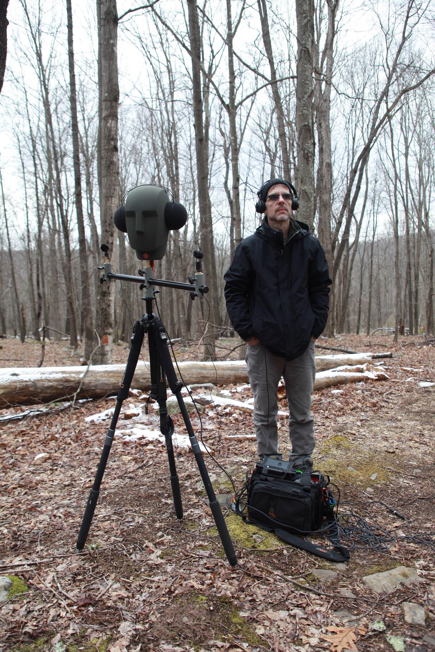 A person stands in a forest with a mannequin head wearing headphones and recording equipment.