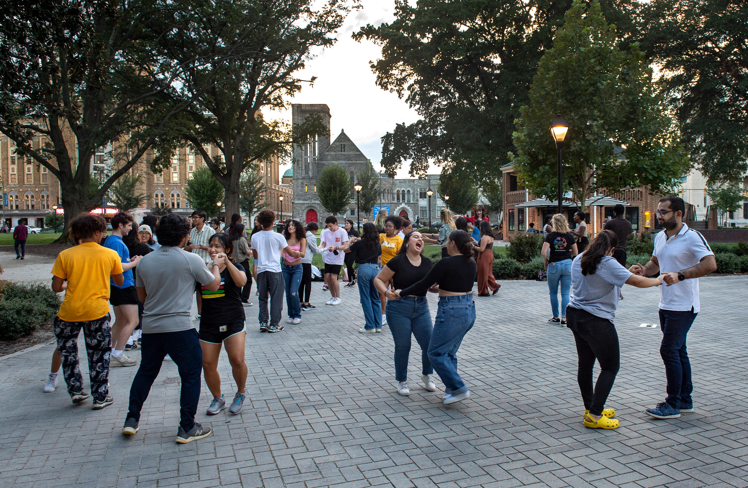 A large group of pairs of students dance on a brick sidewalk lit by the evening sun next to a tree-lined green space in Monroe Park.