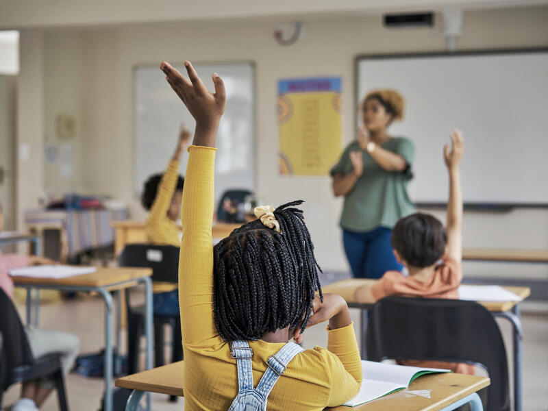 A photo of a classroom of elementary school students raising their hands. In the front of the classroom is a teacher speaking. 