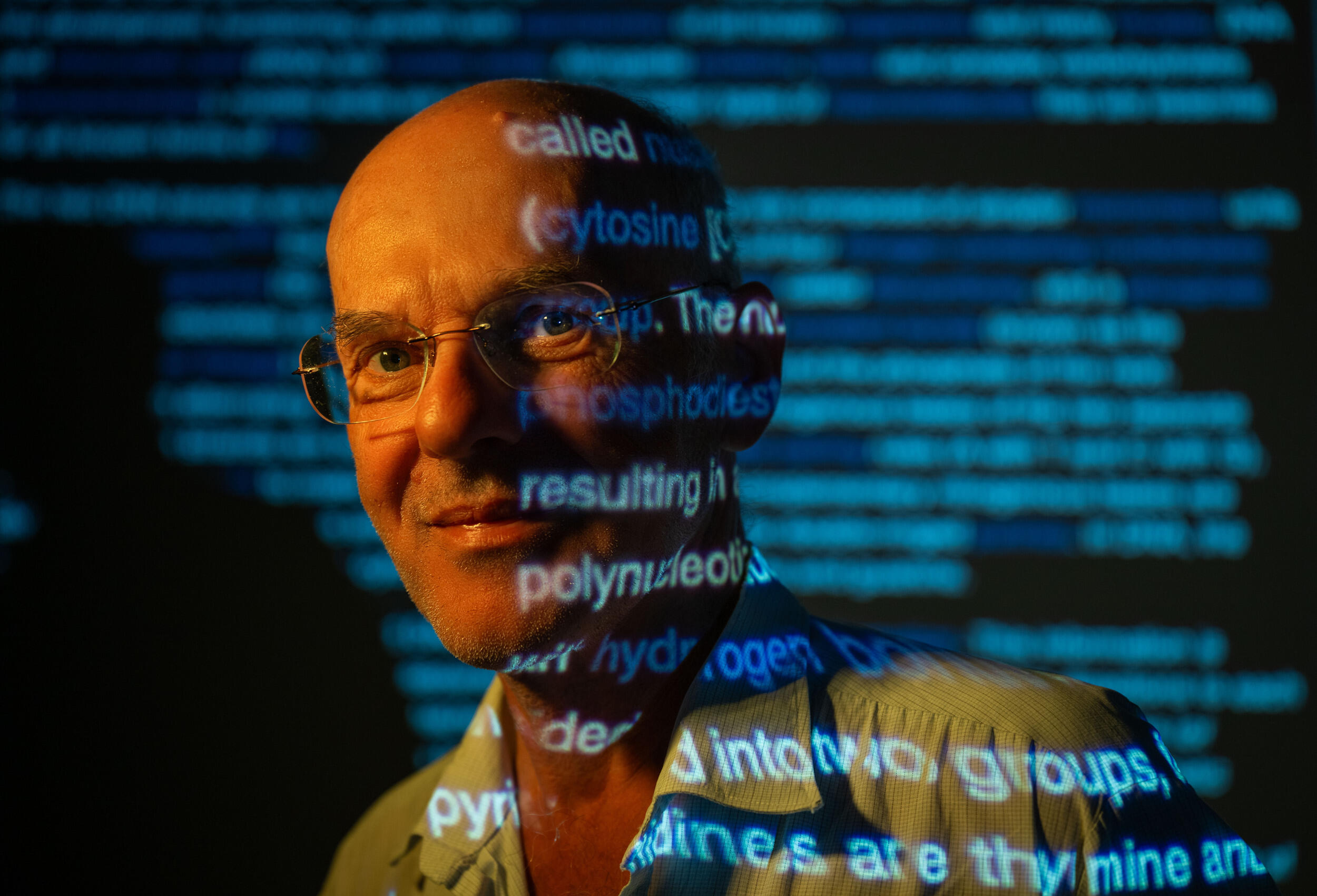 A photo of a man from the shoulders up. There is a projection of blue text that is going across his faace and the wall behind him. 