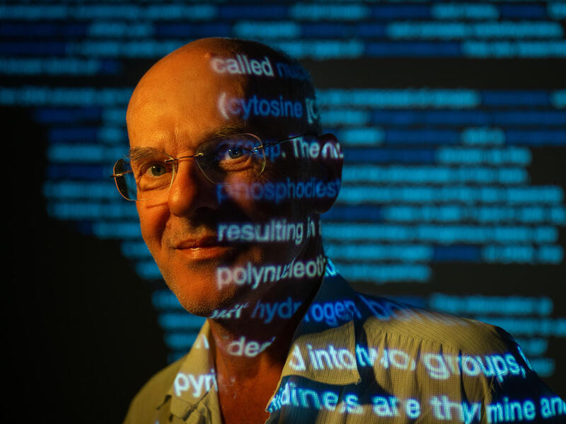 A photo of a man from the shoulders up. There is a projection of blue text that is going across his faace and the wall behind him. 