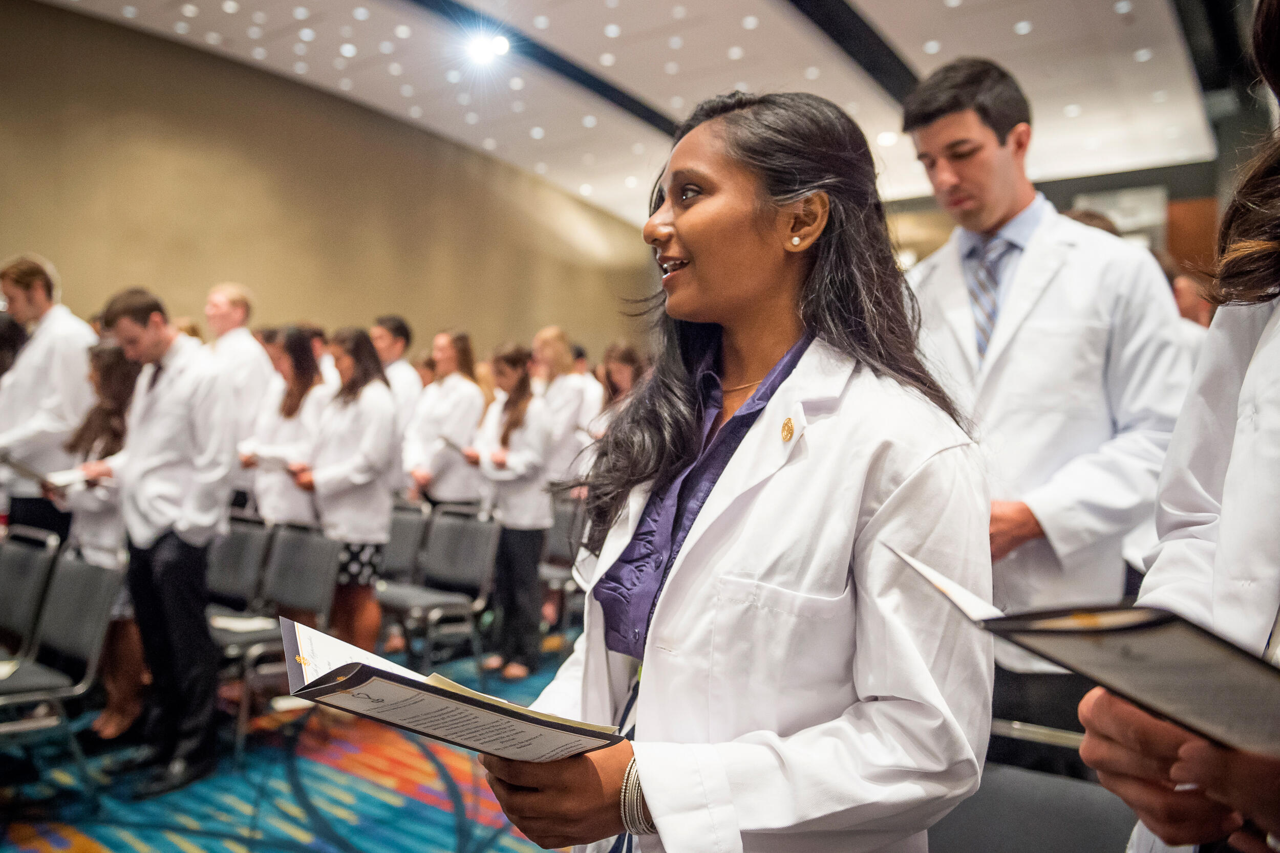 A student in a large auditorium surrounded by fellow students in white medical school coats looks up while reading along with a pamphlet at a ceremony.