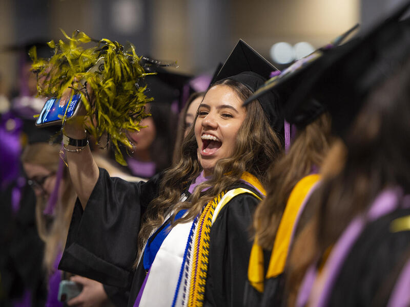A photo of a woman wearing a graduation cap and gown holding a pompom and cheering.She is seated around many other graduating students who are blurry. 