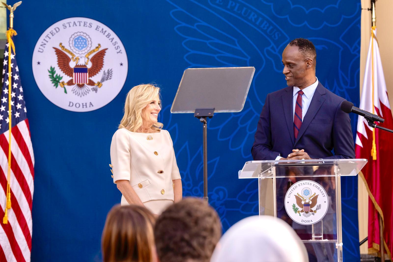 A photo of a man standing at a podium looking at a woman standing to his left. 