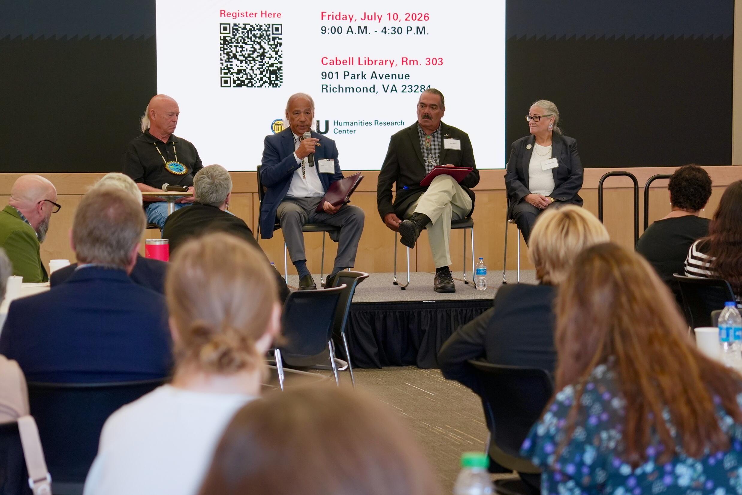A photo of four people sitting on chairs on a stage in front of a crowd of people. 