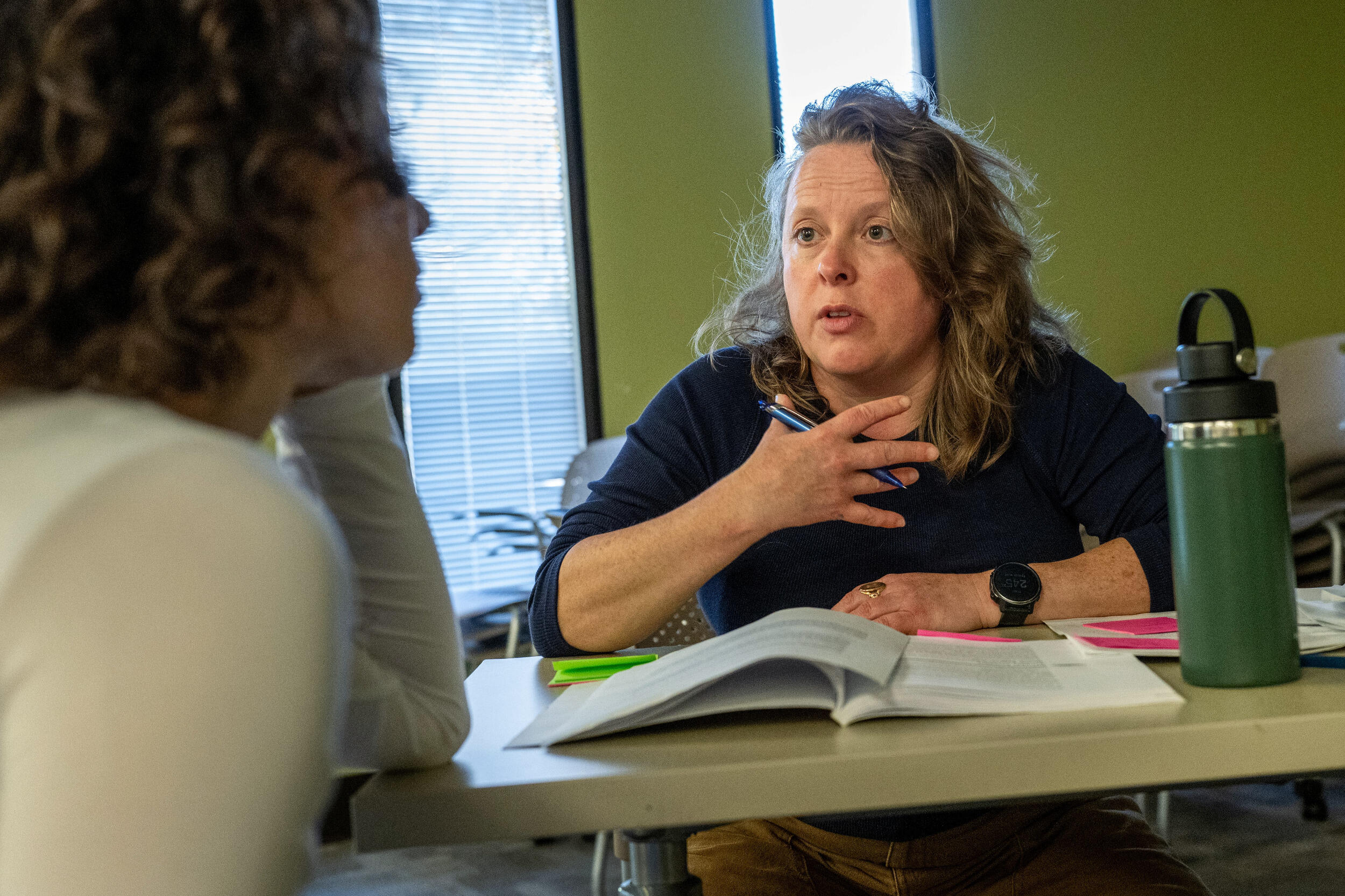 A woman sits at a table and speaks to another woman with an open book in front of her.