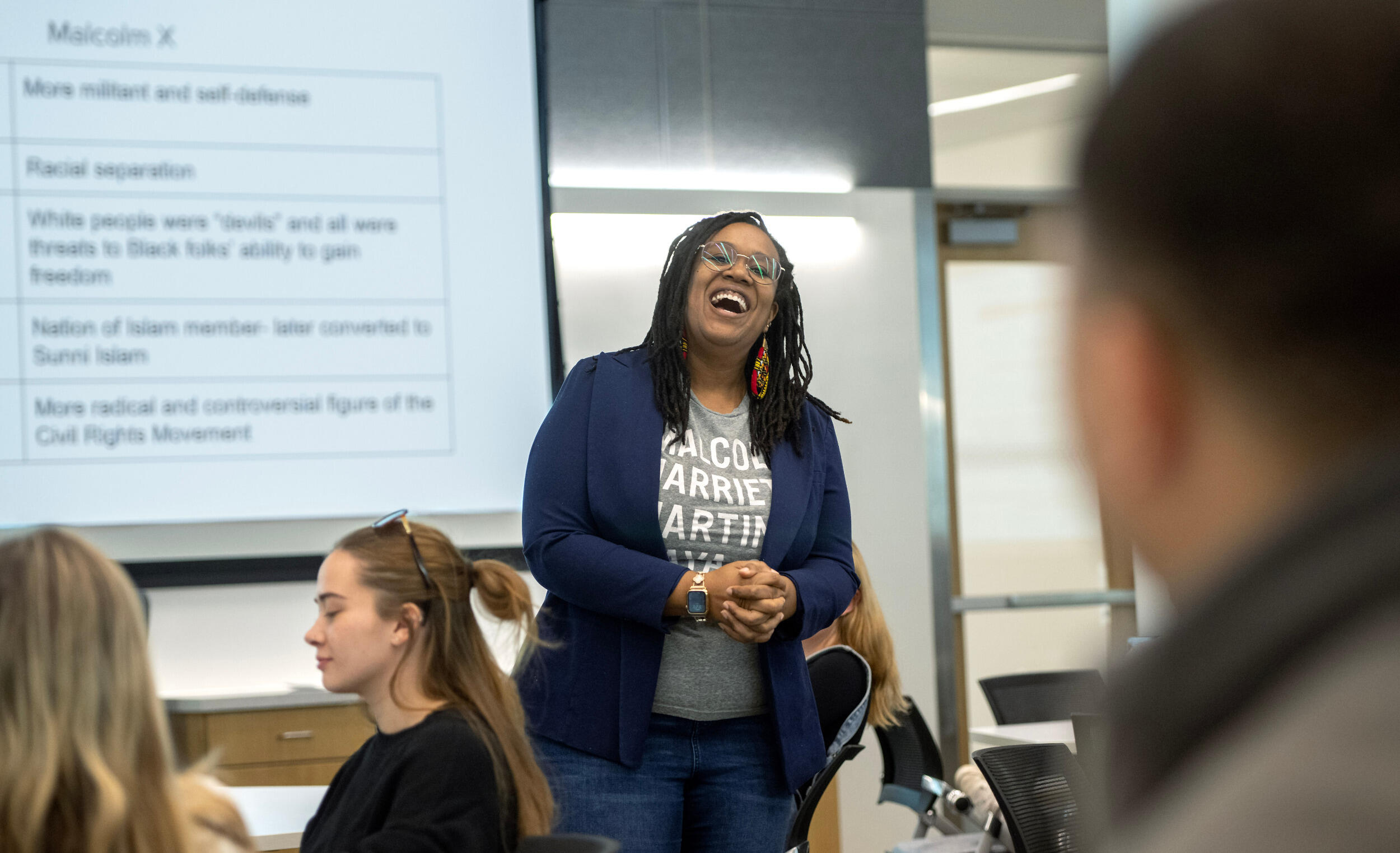 A woman in a T-shirt and blazer laughs standing in front of a screen with text on it.