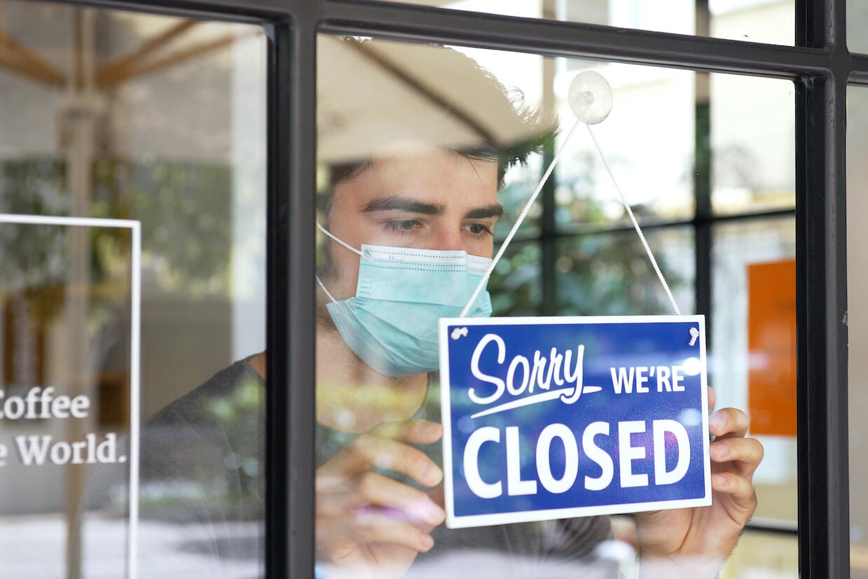 A person wearing a mask holds a \"Sorry we're closed\" sign attached on a window.