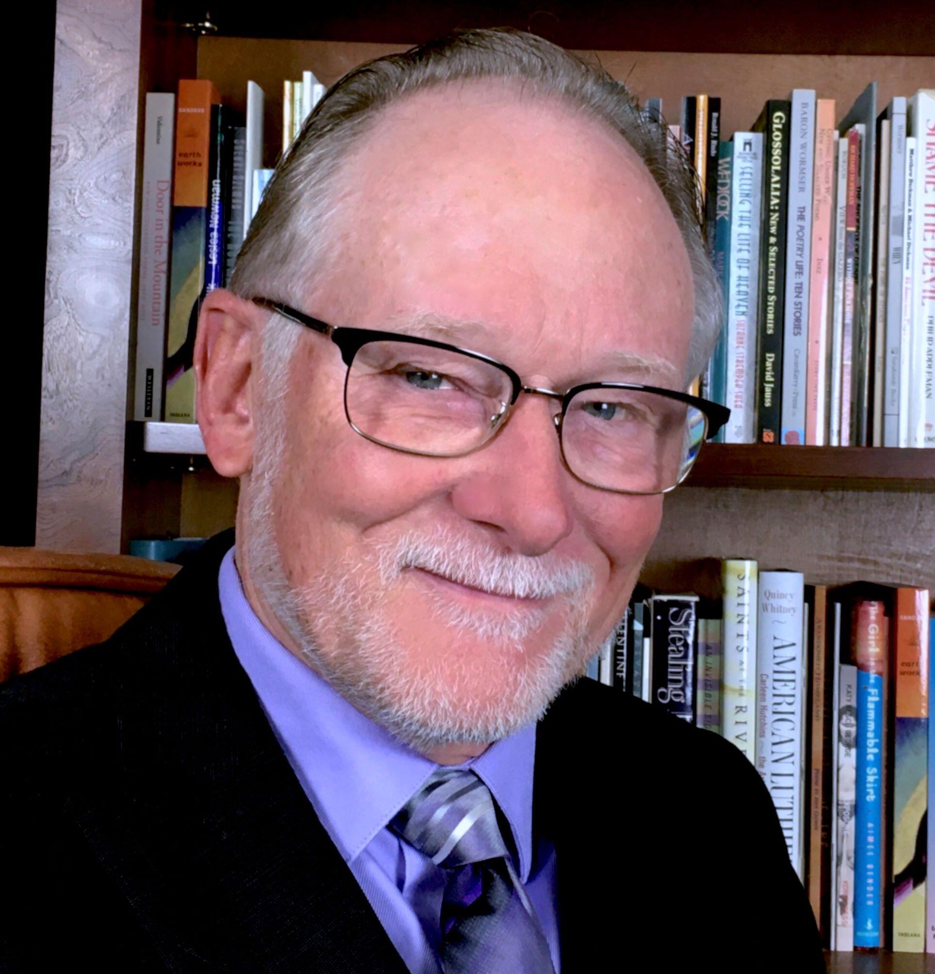 A man in glasses and a coat and tie in front of bookshelves filled with books.