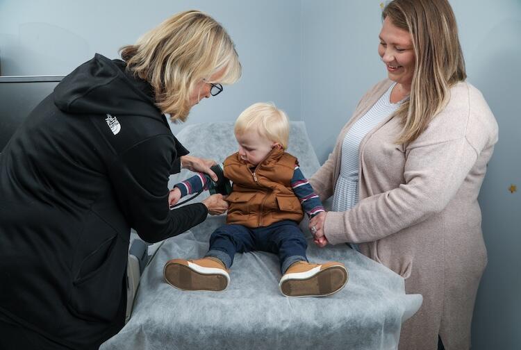 A photo of a todler sitting on a medical examination table. On the left and right of the table are two women looking at the baby. 