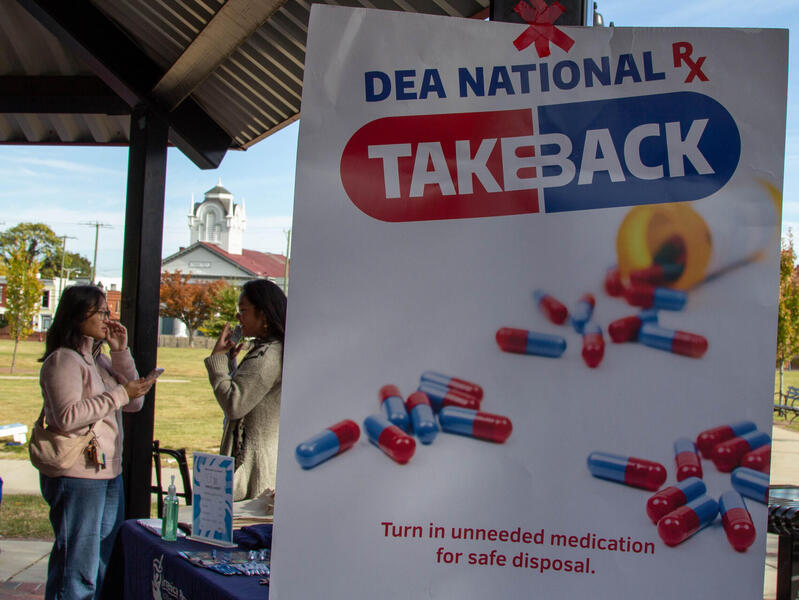 A photo of two women talking to each other next to a table. In front of them is a poster that says \"DEA NATIONAL TAKEBACK\" \"Turn in unneeded medication for safe disposal.\" with a photo of an open pill bottle on it. 