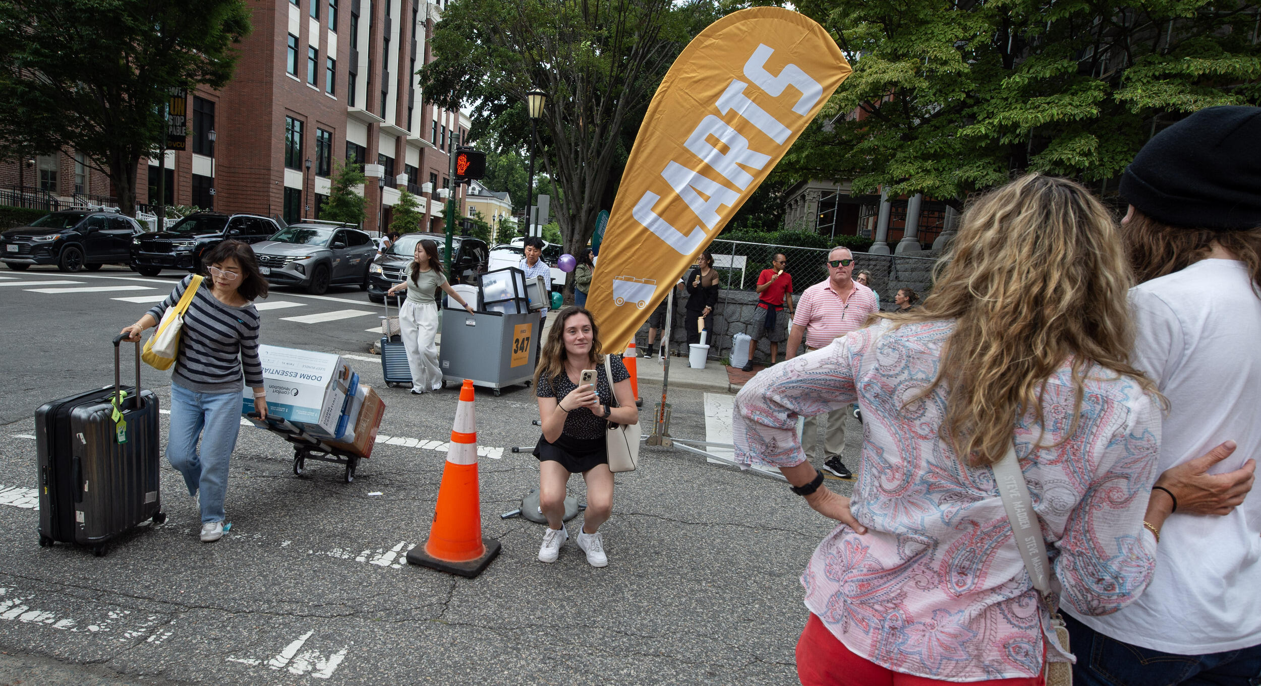 A photo of a street with people walking across it carrying luggage and bins filled with boxes. There is a person taking a photo of two other people. 