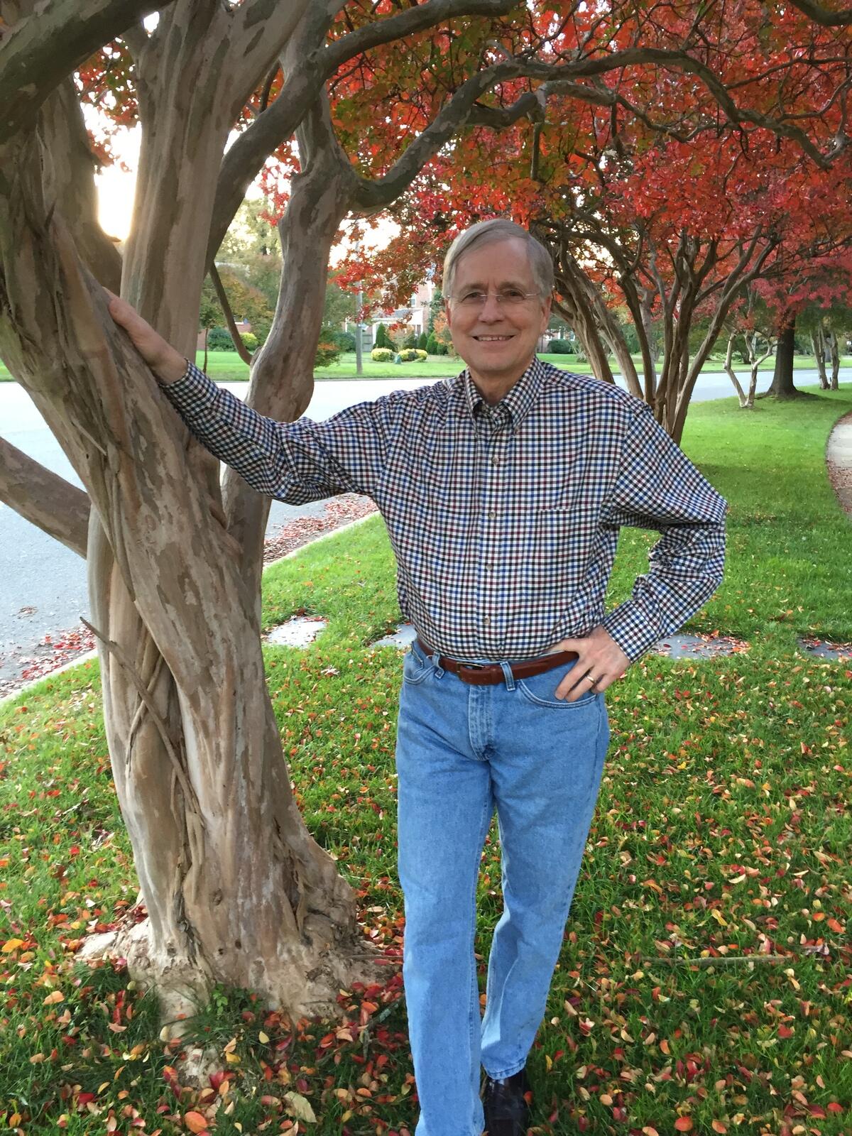 A man with gray hair standing with one hand placed on a tree and the other on his hip. He is wearing a gingham shirt and blue jeans. 
