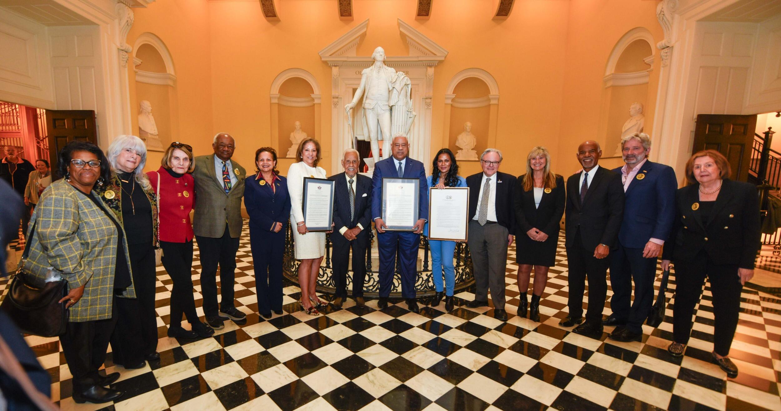 A group photo of 14 people standing in a semi-circle in front of a white marble statue. Three people in the center are holding framed certificates.