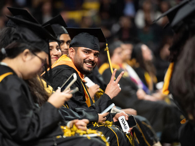 A photo of a row of students sitting at Comencment. THey are all wering graduation cap and gowns. One man in the middle is looking at the camera and putting up a peace sign with his left hand. 