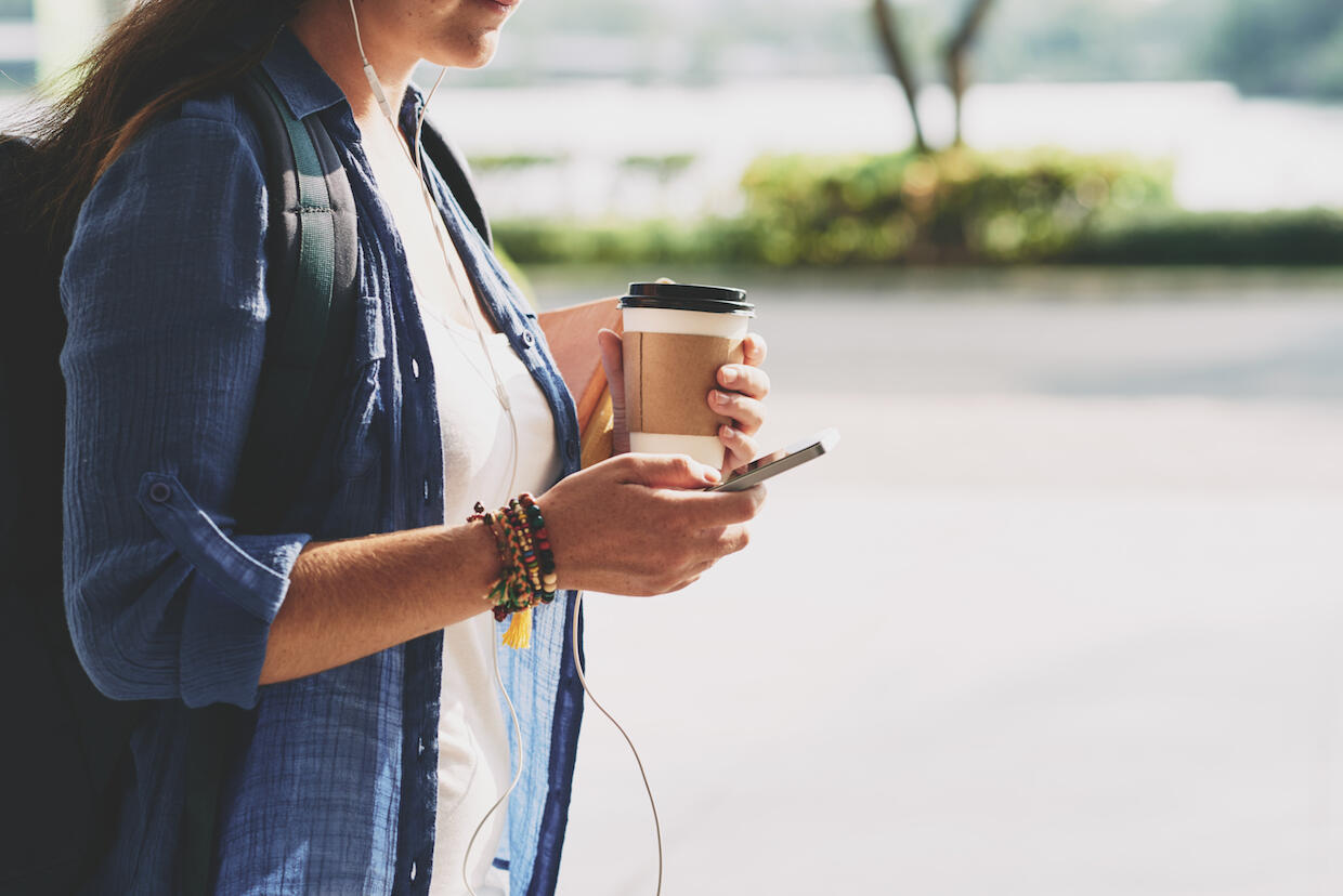 A person wearing headphones and holding a coffee cup and smartphone.