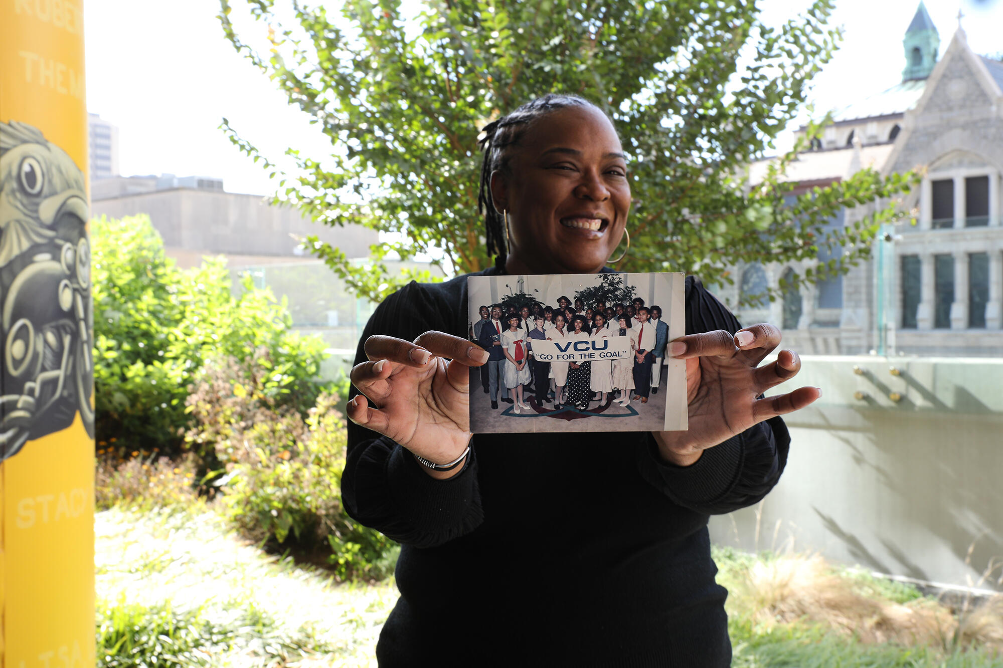 A photo of a woman from the waist up. She is holding a photo in front of her. The photo she is holding shows a group of people standing. In front of the group some people are holding a white banner that says \"VCU Go for the goal\" in black letters. 