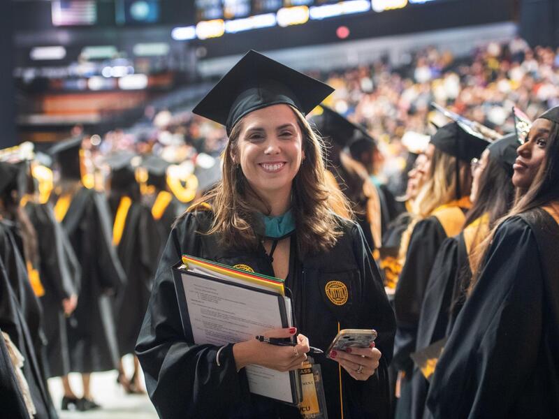 A photo of a woman wearing a cap and gown smiling and standing. In her left hand she has a folder and a binder. In her right hand she is holding a cell phone. All around her is a crowd other people in graduation caps and gowns. 