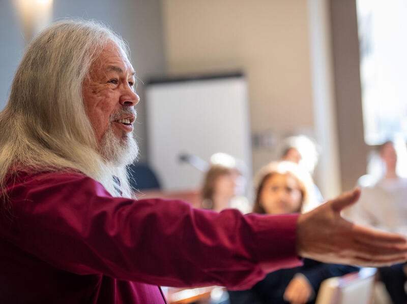 A photo of a man from the chest up. He is speaking to a crowd and extending his right arm to the side. 